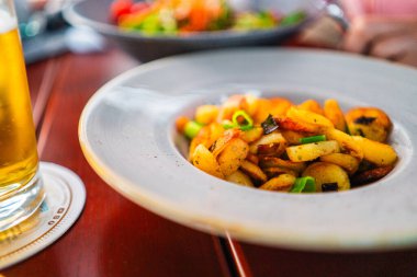 Lezzetli kızarmış patatesler baharatlarla tatlandırılmış ve taze soğanlarla süslenmiş beyaz tabakta. Yakınlarda, ferahlatıcı bir bardak bira yemek deneyimini güçlendirir..