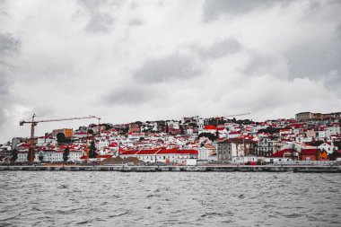 Gri gökyüzüne karşı canlı çatıları olan pitoresk bir kıyı şehri. İnşaat vinçleri, modern gelişim ve tarihsel mimarinin bir karışımını vurguluyor.