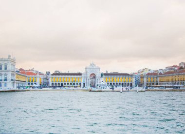 Lizbon 'daki tarihi bir simgenin panoramik görüntüsü, parlak sarı cephelerini ve dekoratif mimarisini gözler önüne seriyor, Tagus Nehri ön planda bulutlu bir gökyüzünün altında..