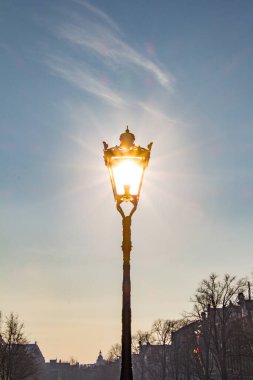 Kaliteli bir sokak lambası, arkasında sabah güneşi ile parlıyor, açık mavi gökyüzüne ve çıplak kış ağaçlarına karşı parlak bir etki yaratıyor..