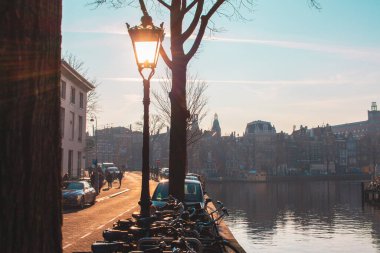 Bicycles and a street lamp line a canal in Amsterdam at sunset, silhouetting historic buildings against a serene, clear blue sky. The tranquil scene evokes a peaceful urban atmosphere. clipart