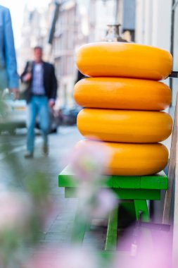 Bright yellow cheese wheels stacked on a green table in a European street. A blurred pedestrian walks by, adding dynamic depth to this urban setting. clipart