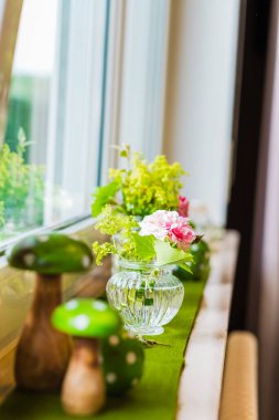 Cam bir vazonun içindeki narin bir çiçek aranjmanı pencere pervazında, yanında da ahşap mantar dekoru bulunur. Sahne doğal ışığı yansıtıyor, dingin ve taze bir ev dekorasyonu atmosferi sunuyor..