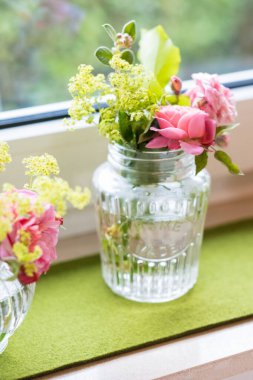 Yeşil bir paspasın üzerinde pembe güller ve sarı çiçeklerin bulunduğu hoş bir çiçek aranjmanı. Güneş ışığıyla aydınlatılmış bir pencere, iç dekorasyon için zarif ve sakin bir atmosfer yaratıyor..