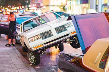 Klasik bir Lowrider arabası, ön tekerleri havada, alacakaranlıkta canlı bir sokak buluşması sergiliyor. Canlı şehir arkaplanı bu otomotiv kültür olayına ekleniyor.