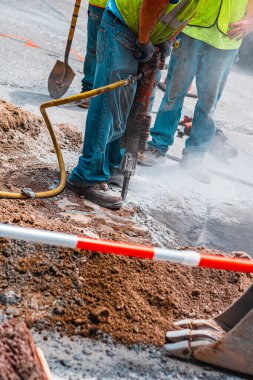 Şehir yolunu kazan, yansıtıcı yelek ve kot giyen, altyapı inşaatına odaklanan, matkaplı işçiler. Güvenlik önlemleri ve engeller aktif, tozlu inşaat alanını gösteriyor