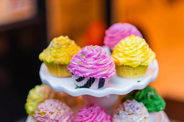 A variety of colorful cupcakes feature pink, yellow, and green frosting on a white stand. Captured in a warm, inviting setting, perfect for festive occasions and celebrations. clipart