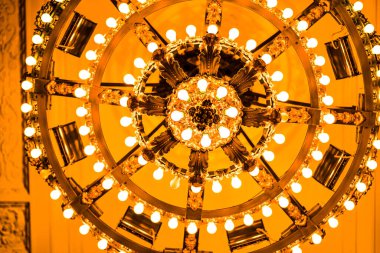 Işıldayan ampullerle dolu büyük bir avizenin ayrıntılı görüntüsü, süslü altın detaylar sergileniyor. Ortamın sıcaklığı ve zenginliği barok tasarımının iç dekordaki zarafetini vurguluyor.