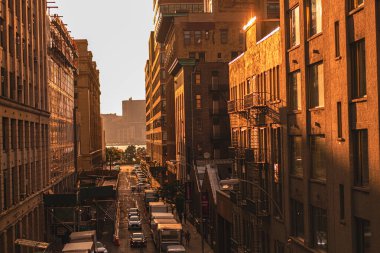 Altın saatinde hareketli bir şehir caddesi, güneş ışığı tuğla binalara sıcak bir ışık saçıyor. Sokaklar arabalarla dolu ve arka planda manzaralı bir nehir görünüyor..