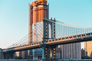 Görüntü, Manhattan Köprüsü ve New York 'ta gün batımında inşa halindeki bir gökdelenin kent mimarisini ve şehrin canlı şehir manzarasını gözler önüne seriyor..