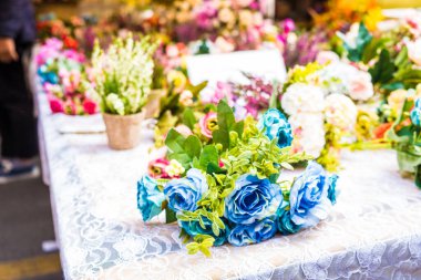 Mavi güller ve canlı yapraklardan oluşan güzel bir düzenleme dantelli bir masanın üzerinde duruyor. Renkli çiçeklerle çevrili ekran, pazarda davetkar ve canlı bir atmosfer yaratıyor..