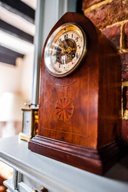 Kaliteli ahşap bir saat, sıcak ve davetkar bir odadaki tuğla duvarla örülmüş cilalı bir şömine rafına ustalıkla yerleştirilir. Saatin antika tasarımı zamansız işçiliği vurguluyor..