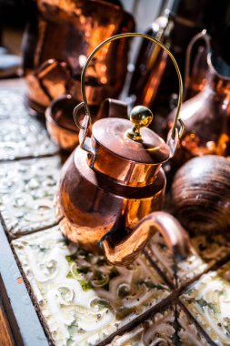 Antika bir bakır çaydanlık ve antika yemek takımı dekoratif fayanslarla döşenmiş bir yüzeyde, sıcak tonlarda ve karmaşık yansımalarla dinleniyor. Kırsal bir mutfağın cazibesini ve nostaljisini mükemmel bir şekilde yansıtıyor..