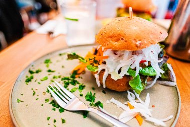 Paramparça sebzeler ve yapraklı yeşillikler gibi taze malzemeler içeren vejetaryen bir hamburgerin canlı bir yakın çekimi gündelik bir yemek ortamında maydanoz garnitürüyle şık bir tabakta sunuluyor..