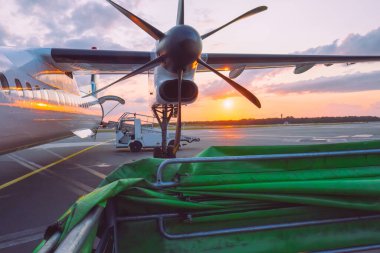 A propeller plane sits on the runway at sunset, casting warm reflections on its fuselage. Nearby, cargo equipment waits for loading. The scene exudes a sense of travel and adventure. clipart