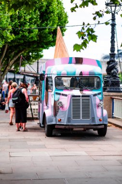 Yapraklı ağaçların altında hareketli bir caddeye park etmiş renkli bir dondurma kamyonu sıcak ve güneşli bir günde lezzetli şeylerden zevk almak isteyen insanları cezbediyor. Yaz ve sokak yiyeceklerini yakalar..