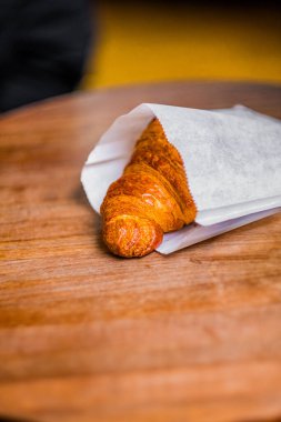 Yeni pişmiş bir kruvasan, beyaz bir kağıda sarılmış, ahşap bir masada oturur. Altın, pullu hamur işi görsel olarak çekici, onu kahvaltı için mükemmel yapıyor ya da hızlı bir aperatif olarak.