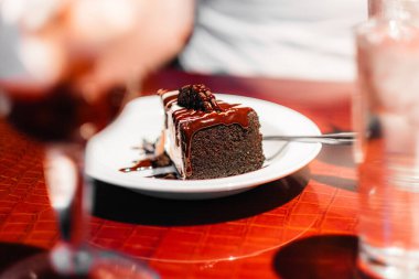 Beyaz bir tabakta servis edilen parlak çikolata sosuyla kaplı yozlaşmış bir dilim çikolatalı pasta. Kafenin sıcacık havası bu tatlı deneyimine sıcak, davetkar bir dokunuş katıyor..