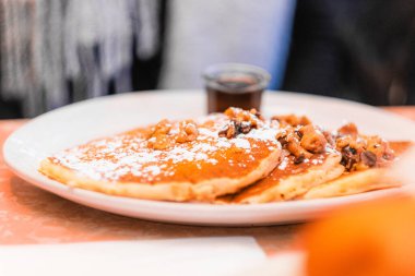 A close-up of fluffy golden pancakes topped with walnuts and powdered sugar, accompanied by a side of syrup. Perfect for a warm, inviting breakfast or brunch setting. clipart