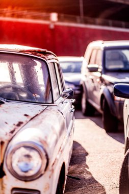 Bu resim, güneşli bir otoparkta, eski ve yeni ulaşım araçları arasındaki zıtlığı gösteren, paslı bir dış görünüşü olan klasik bir arabayı gösteriyor..