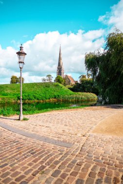 Büyüleyici bir kilise kulesi, parlak mavi bir gökyüzüne karşı uzanan parke taşı yolu ve huzurlu bir nehrin yanında yemyeşil bir arazinin üzerinde yükseliyor. Huzurlu ve manzaralı temalar için mükemmel.