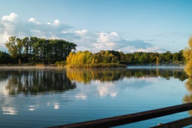 Sakin bir göl yemyeşil bir orman ve yumuşak bir sabah sisi yansıtır, berrak mavi gökyüzüne karşı huzurlu ve sakinleştirici bir atmosfer yaratır. Doğa ve rahatlama temaları için ideal.