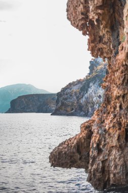 Sakin bir deniz ve berrak bir gökyüzü karşısında, arka planda uzak dağlarla sarp kıyı kayalıklarının nefes kesici manzarası. Huzuru, doğal güzelliği ve macera duygusunu aktarmak için idealdir..