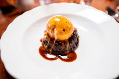 Zengin karamel sosu ve kremalı dondurmayla süslenmiş bu çikolatalı lav keki ile mükemmel bir tatlı ziyafeti için zarif beyaz bir tabakta servis edilen bir gurme deneyimi yaşayın..