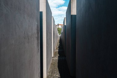 Yüksek beton duvarlarla çevrili dar bir yol, parlak ışıklı binalara doğru uzanır. Mavi gökyüzü ve kabarık bulutlarla çarpıcı bir kontrast yaratır..