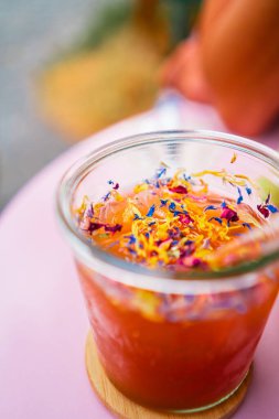 Cam bir kavanozdaki ferahlatıcı bir içeceğin üzerinde renkli çiçek süsleri, ahşap bir bardak altlığı üzerine kurulmuş, canlı bir yaz gününün özünü yakalıyor. Açık hava eğlencesi ve şenlik için mükemmel..