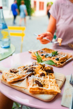 Şurup serpilmiş ve naneyle süslenmiş Belçika wafflelarının yakın çekimi, açık bir kafede servis ediliyor. Rahatlatıcı bir atmosferde sakin bir kahvaltı ya da brunch için hoş bir ortam..