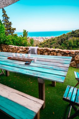 Yıpranmış ahşap mobilyalarla, okyanus manzarası, şehir manzarası ve mavi gökyüzünün altındaki ağaçlık tepelere bakan taşralı oturma düzeniyle. Tatil, rahatlama ve doğa temaları için.