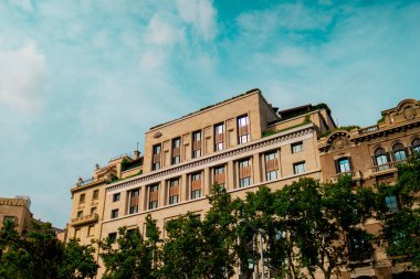 Başkent ortamında, yemyeşil ağaçlarla çevrili ve mavi gökyüzü olan açık bir günde resmedilen klasik bir bina..