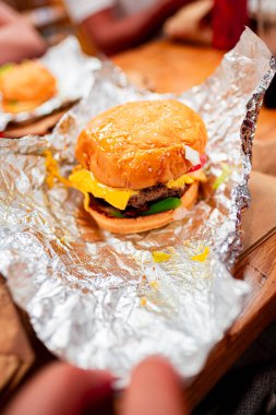 Taze malzemeler ve erimiş peynirli, kısmen folyoya sarılı, ahşap bir masada sıcak ve lezzetli bir yemek deneyimi yakalayan bir çizburgerin yakın çekimi..