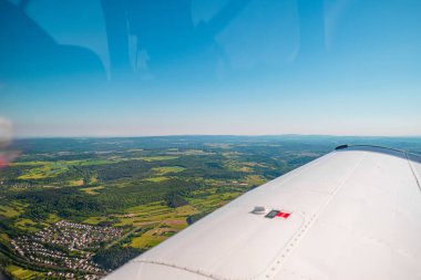 Açık alanları ve doğal güzelliği vurgulayan açık mavi gökyüzünün altında, bir uçak kanadından görülebilen orman ve tarlalarla birlikte kırsal bir arazinin manzaralı görüntüsü..