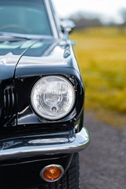 Klasik siyah arabanın ön farları doğal ışık altında, zarif tasarımı ve zamansız cazibesiyle sergileniyor. Parlak, bulanık arkaplan arabanın çarpıcı görünümünü tamamlıyor.