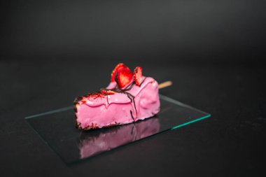 Şişte pembe çilekli kek dilimi, çikolata serpiştirilmiş ve taze dilimlenmiş çilekle süslenmiş, cam bir tabakta dinleniyor.