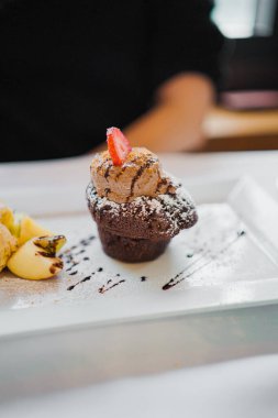 Çilek dilimiyle süslenmiş iştah açıcı çikolatalı bir tatlının yakın çekimi, beyaz bir tabakta sunuluyor, vanilyalı dondurma ve çikolata soslu bir sunum sunuluyor..