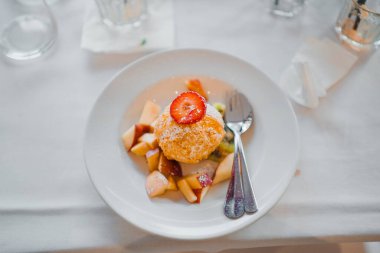 Üzerinde çilek dilimi olan ve beyaz bir tabakta taze meyve parçalarıyla servis edilen altın kızartılmış bir hamur tatlısı sunumu gündelik bir ambiyansla bir masada sunuluyor..