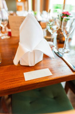 An elegant dining table setup in a restaurant or banquet hall, showcasing a crafted white folded napkin placed alongside a name card, glassware, and utensils on a polished wooden table. clipart