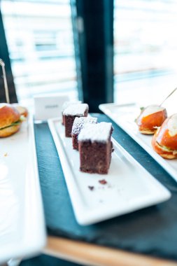 Pudra şekerli çikolatalı browniler şık beyaz bir tabakta sunuluyor, sandviçlerin yanında, etkinlikler veya gelişigüzel toplantılar için davetkar bir görüntü yaratıyor..