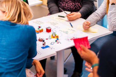 A group of people engage in a collaborative brainstorming session, using Lego models, handwritten notes, and discussions to develop ideas and solutions in a creative and engaging manner. clipart