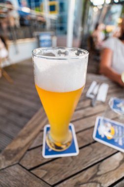 Açık hava ortamında ahşap bir masada servis edilen ferahlatıcı uzun bir bardak köpüklü bira. Rahatlamak ve sosyalleşmek için ideal bir ortam..