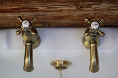 Konforlu Banyo 'daki klasik pirinç musluklar: Sıcak ahşap zemine karşı pirinçten muslukları olan rahat, kırsal bir banyo lavabosu. Eski tarz musluklardan birinden su akmaktadır. Pirinç ve ahşap karışımı banyoya eski moda bir cazibe katar..