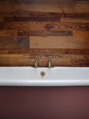 Pembe seramik bir küvete yakın plan pirinç musluklar, ahşap bir panel duvara dayanıyor. Bu resim, banyo dekoru, iç tasarım ilhamı veya klasik temalı alanlar için idealdir..
