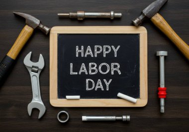 Çekiç, ingiliz anahtarı ve metal parçaları gibi çeşitli aletlerin yer aldığı ve 'İşçi Bayramı' mesajını gösteren bir karatahtanın etrafına yerleştirilmiş durgun bir yaşam kompozisyonu. Sahne, karanlık, ahşap bir yüzeye göre ayarlanmış.