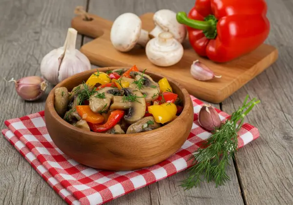 Kırsal bir tabak saut mantarı ve renkli dolma biber kareli bir peçete üzerinde ahşap bir kasede sunulur. Taze sarımsak karanfilleri, dereotu filizi ve mantarlı ahşap kesme tahtası ve kırmızı dolma biber mutfak sahnesine ilave ediliyor..