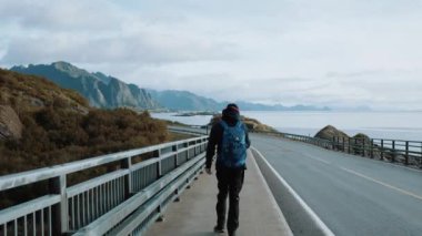 Genç adam Lofoten adalarındaki dağ ve deniz manzaralı yolda yürüyor.