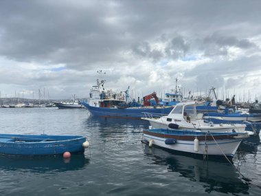 Parlak bir Akdeniz gökyüzü altında tarihi yapılar tarafından çerçevelenmiş, berrak mavi sularda sallanan renkli tekneleriyle canlı Ischia limanı.