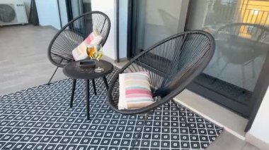 Balkonda iki sandalye, elinde iki kadeh bardakla duran bir masa. Dönen bir manzarada çekilmiş. Rahatlatılmış açık hava eğlencesi.